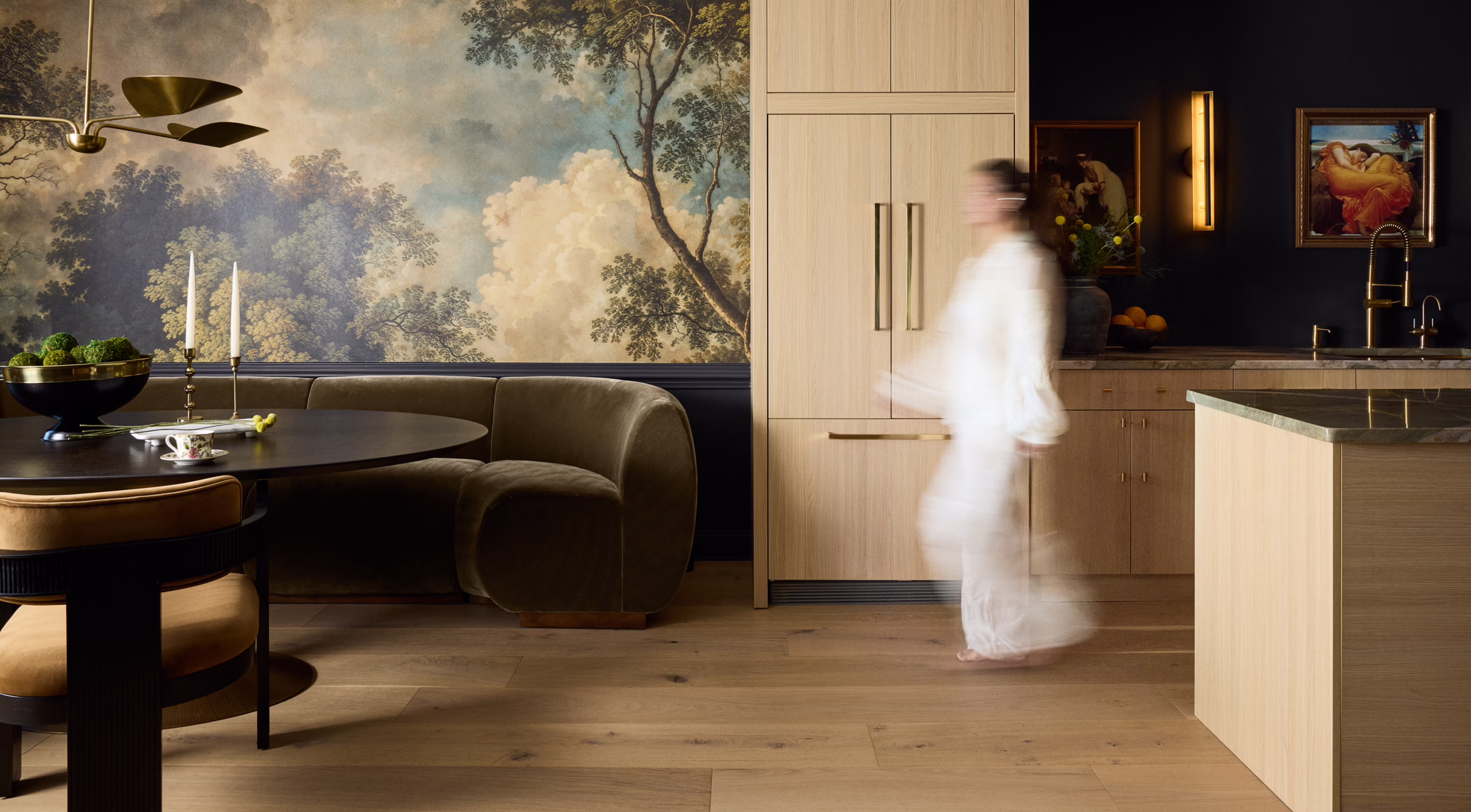 Woman walking through a kitchen and dining area with light wood cabinetry and a landscape mural