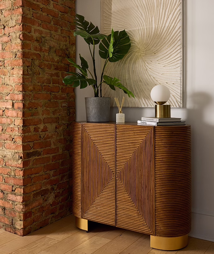 Sculptural wood cabinet beside an exposed brick wall and potted plant